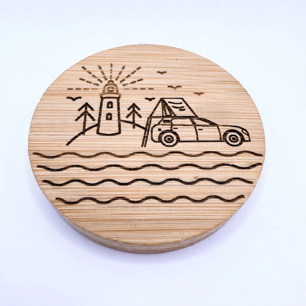Gebranntes Küstenmotiv auf runder Bambusuntersetzer mit Leuchtturm, Bäumen, Auto mit Dachzelt und Wellen.